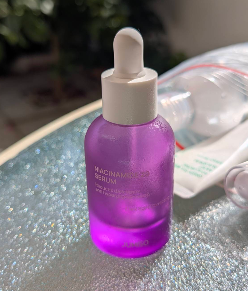 Em uma mesa de vidro, iluminada pelo sol está um potinho de vidro roxo (lindo) com uma tampa conta-gotas branco. O vidro é estilo jateado e a transparência dá um efeito lindo nos tons de roxo.